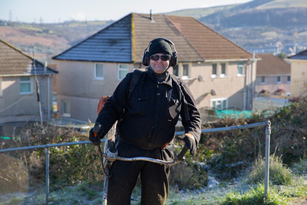 Trivallis Housing Landlord Wales A person stands outside holding landscaping equipment, dressed in dark work attire, protective gloves, and earmuffs. They are in front of houses with hills in the background, with a metal railing nearby.