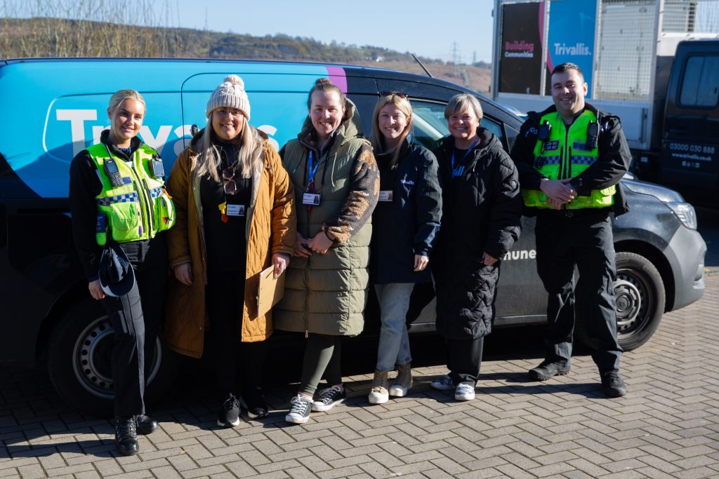 Trivallis Housing Landlord Wales A group of six people stand in front of a black van with "Trivallis" written on the side. Four people wear jackets and lanyards, while two wear police uniforms with high-visibility vests. They are all smiling and standing on a paved surface.