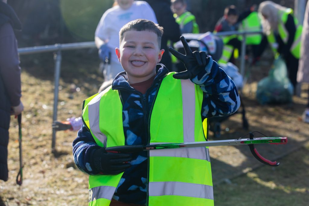 Trivallis Housing Landlord Wales A young boy in a blue and black jacket, wearing a bright yellow safety vest, smiles and flashes a peace sign. He holds a litter picker. Other people in similar vests are in the background participating in a cleanup activity.