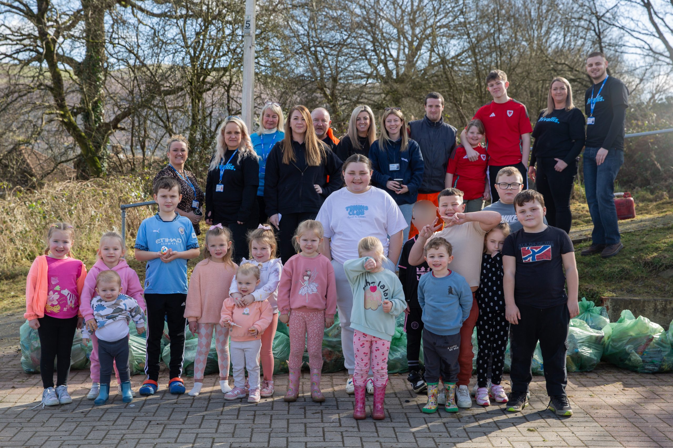 Trivallis Housing Landlord Wales A group photo of adults and children standing outdoors on a paved area. Some children are holding hands; others have their arms crossed. Behind them are numerous green garbage bags. Trees and bushes are visible in the background.