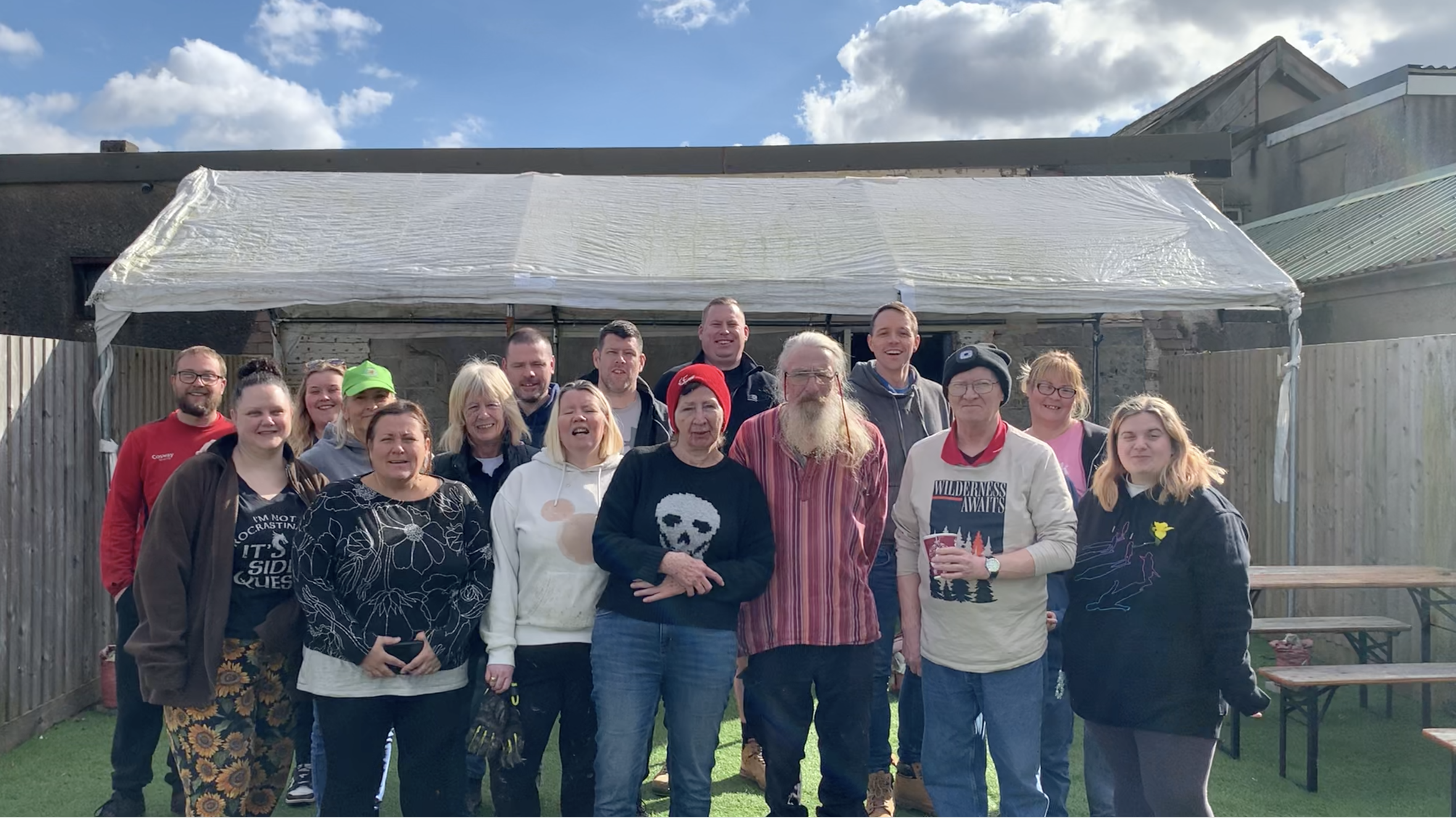 Trivallis Housing Landlord Wales A group of fifteen adults stand together outdoors in front of a rustic wooden structure. The sky is partly cloudy. Some people are smiling, and various clothing styles are visible. A couple of benches are off to the side.