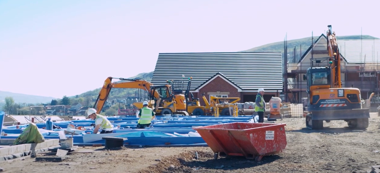 Trivallis Housing Landlord Wales Construction workers in safety vests work on a building site with blue materials on the ground, an excavator, machinery, and a skip. A brick house with a striped roof and scaffolding is visible in the background.