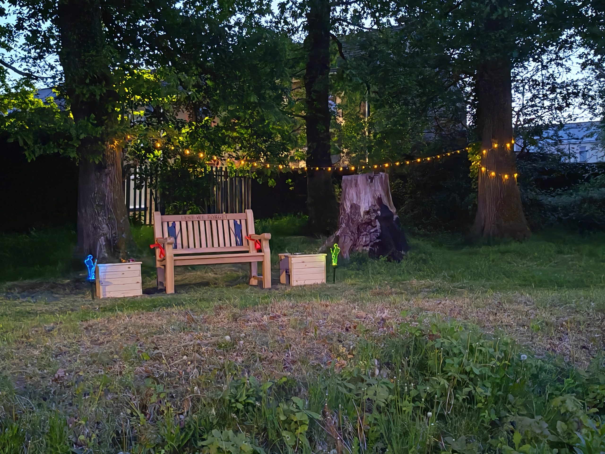 Trivallis Housing Landlord Wales A wooden bench is placed between two large trees in a grassy area, flanked by two wooden boxes. String lights hang between the trees, with a tree stump visible nearby. The scene is outdoors in the evening.