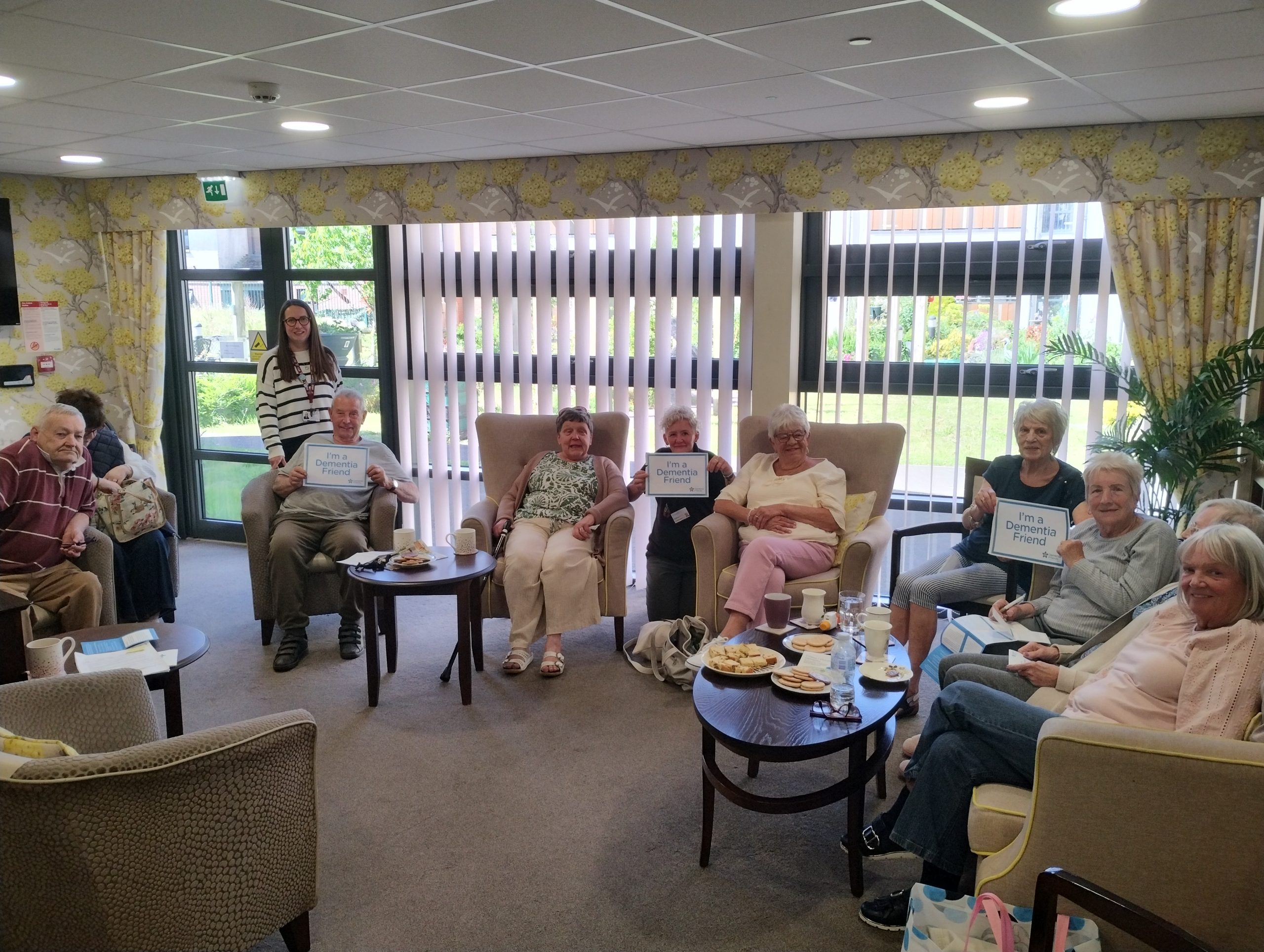 Trivallis Housing Landlord Wales A group of older adults sits in armchairs around tables with food and drinks in a well-lit room. Some are holding signs, and a younger woman stands at the back. Large windows with vertical blinds are behind them.