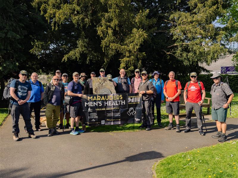 Trivallis Housing Landlord Wales A group of men stand outdoors on a sunny day, holding a banner that reads "Marauders Men's Health." Trees and grass are visible in the background. The men are wearing casual and hiking clothing.