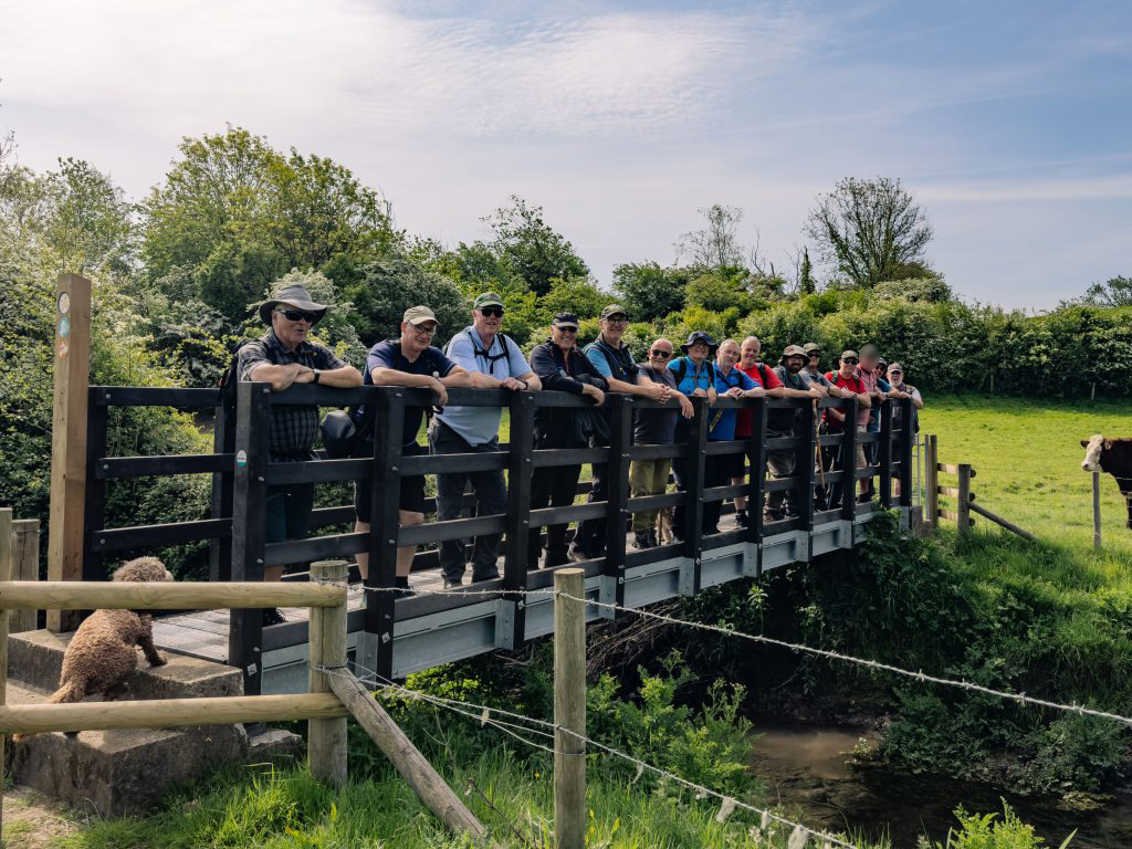 Trivallis Housing Landlord Wales A group of people stand side by side on a small footbridge over a stream, surrounded by greenery. A dog sits nearby on the left, and a cow is partially visible on the right. The sky is partly cloudy.