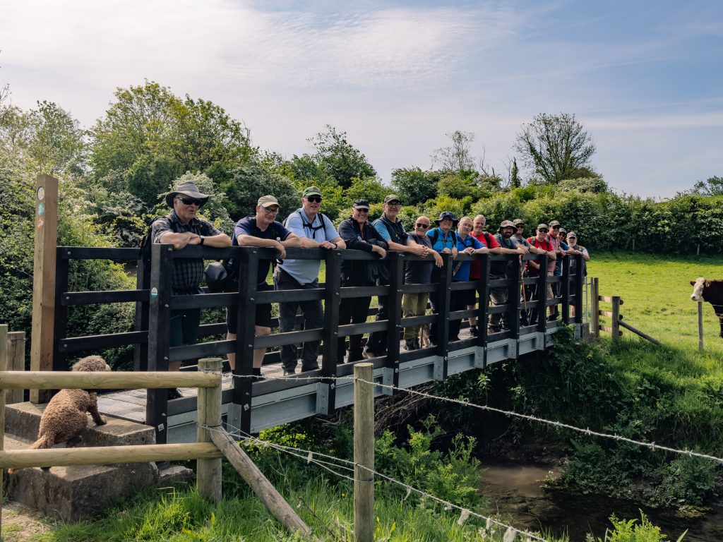 Trivallis Housing Landlord Wales A group of around sixteen people stands side by side on a small wooden bridge over a stream, with trees and bushes in the background. A dog sits on the grassy bank near the bridge. The weather is sunny.