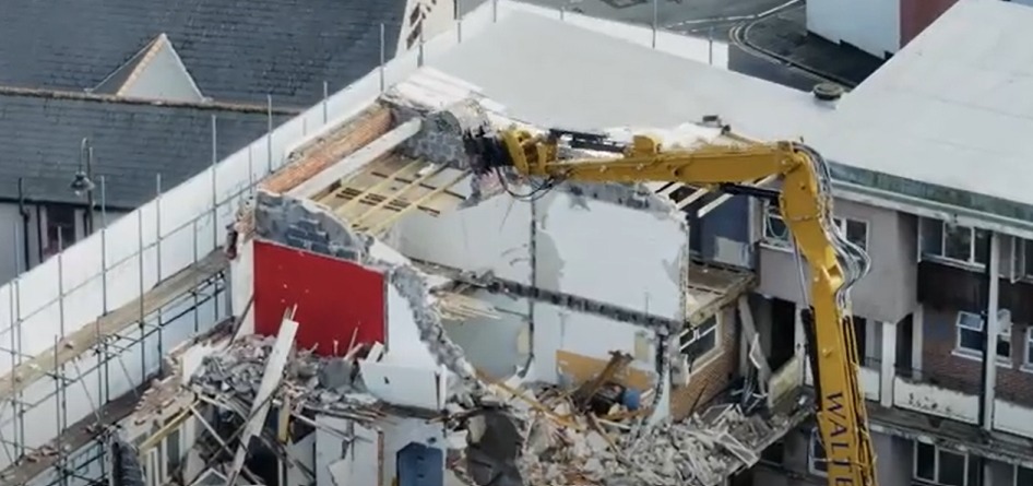 Trivallis Housing Landlord Wales A yellow excavator demolishes the upper floor of Mitchell Court,a partially destroyed building, with debris scattered around and adjacent structures visible in the background.