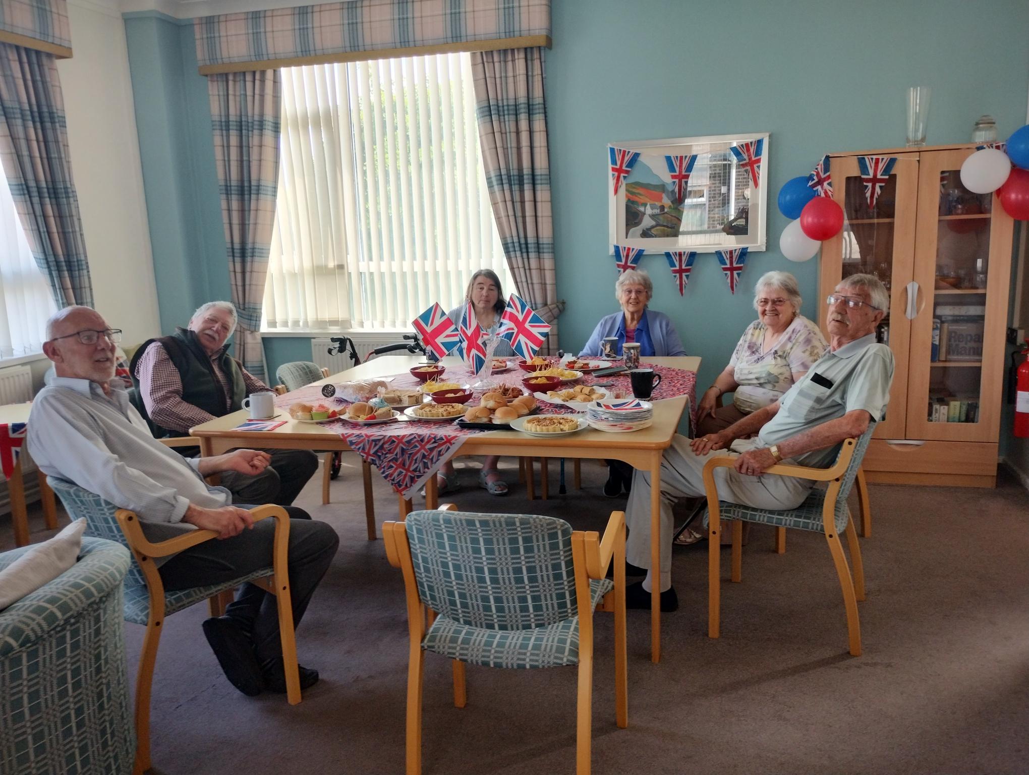 Trivallis Housing Landlord Wales Six older adults sit around a table filled with food, decorated with Union Jack flags. The room has large windows, blue walls, balloons, and additional flags. The atmosphere appears festive and celebratory.