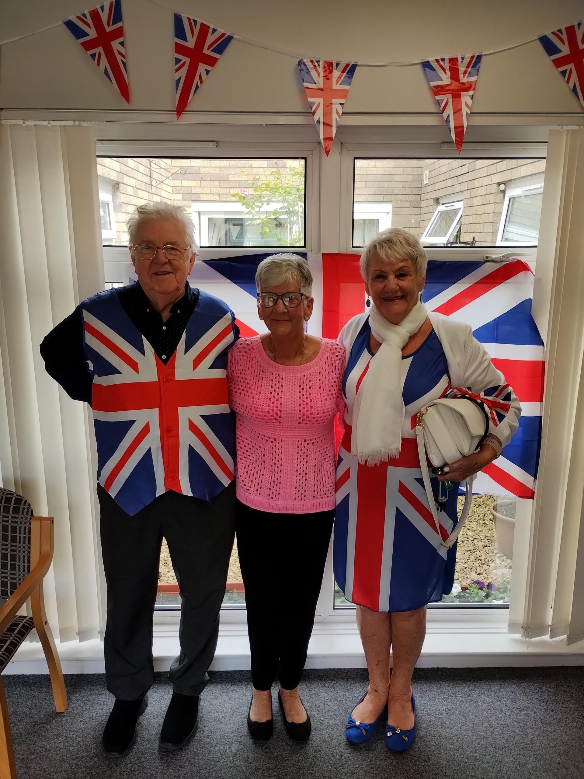 Trivallis Housing Landlord Wales Three older adults stand indoors in front of a window with a Union Jack flag and bunting. Two wear Union Jack-themed clothing, and one wears a pink top. They are smiling and standing close together.