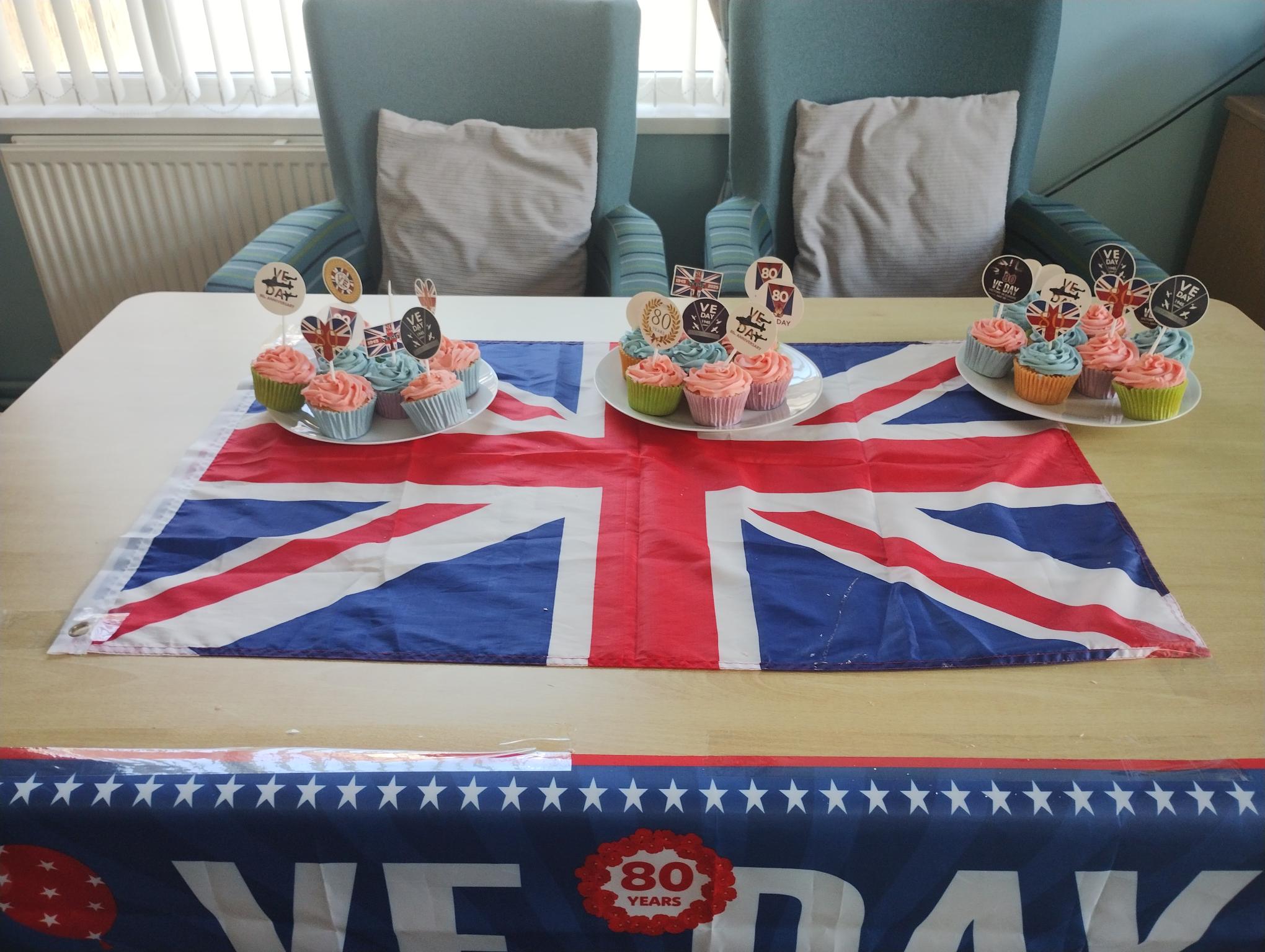 Trivallis Housing Landlord Wales A table with a Union Jack flag, decorated with trays of cupcakes topped with themed toppers. Two armchairs are in the background, and a VE Day banner with "80 years" is draped across the table edge.