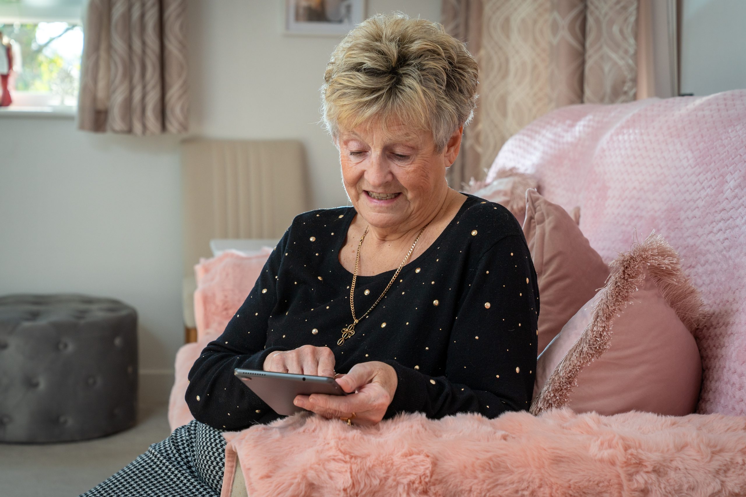 Trivallis Housing Landlord Wales An older woman with short blonde hair sits on a pink sofa, using a tablet. She is wearing a black, long-sleeved top with gold dots and a necklace. The setting appears to be a cozy, well-lit living room.