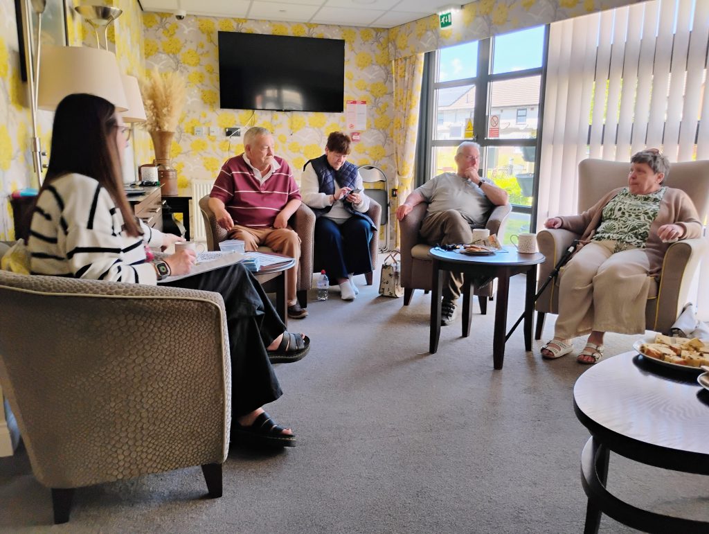 Trivallis Housing Landlord Wales Five older adults sit in a living room, some with tea and snacks on tables. They are engaged in conversation; sunlight comes in through large windows with vertical blinds. The room has floral wallpaper and a mounted TV.