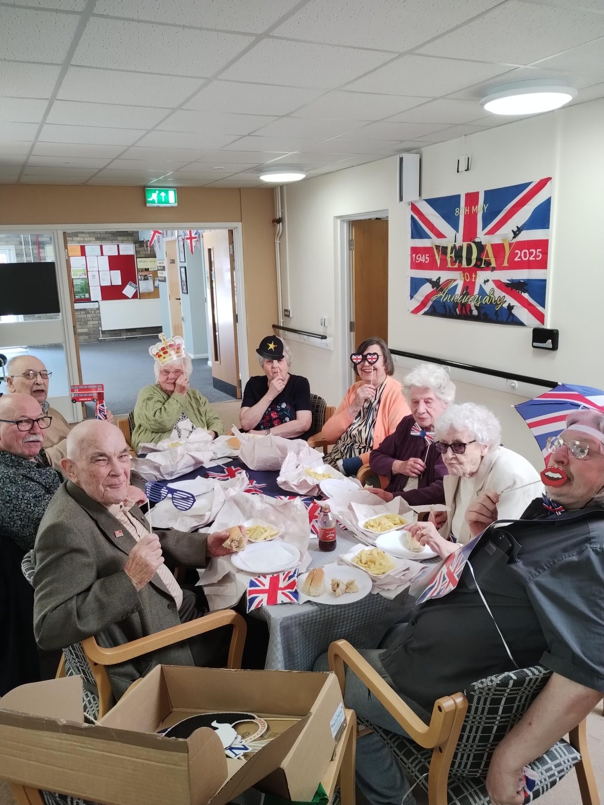 Trivallis Housing Landlord Wales A group of elderly people sit around a table eating food and smiling. The room is decorated with Union Jack flags and a VE Day banner on the wall. Some people wear hats and glasses as accessories.