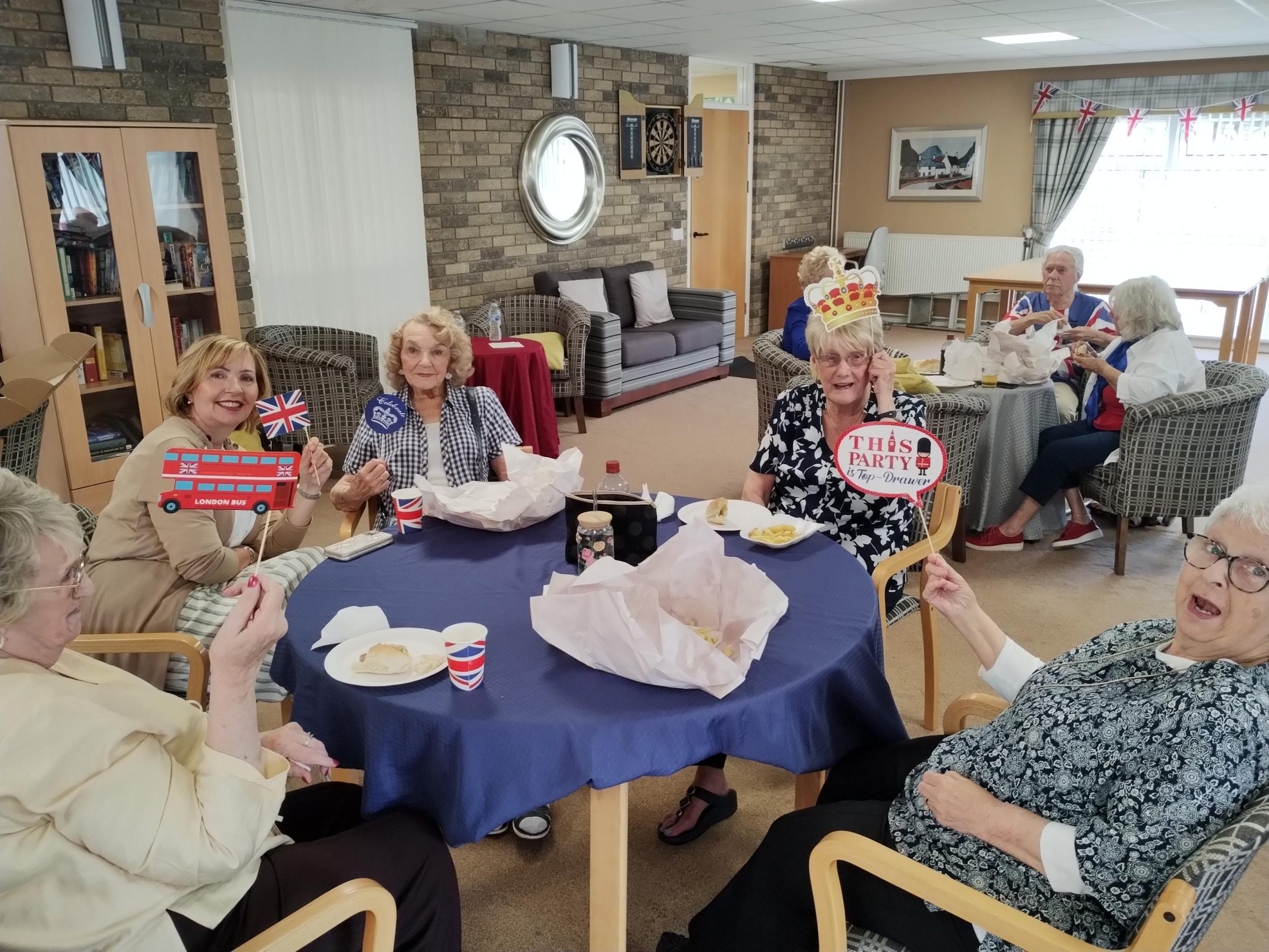 Trivallis Housing Landlord Wales A group of elderly women sit around a table with food and drinks, holding British-themed props and signs. The room has armchairs, decorations, and a dartboard on the wall, giving a festive atmosphere.