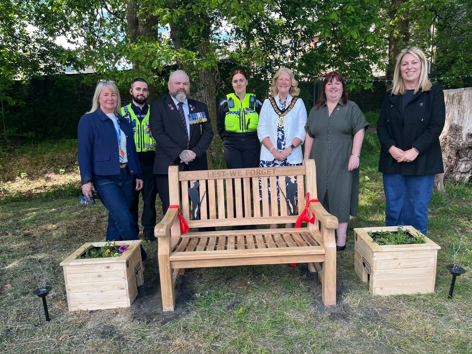 Trivallis Housing Landlord Wales Seven people stand behind a wooden memorial bench inscribed with "LEST WE FORGET," flanked by two planter boxes with flowers, in an outdoor setting with grass, trees, and sunlight.