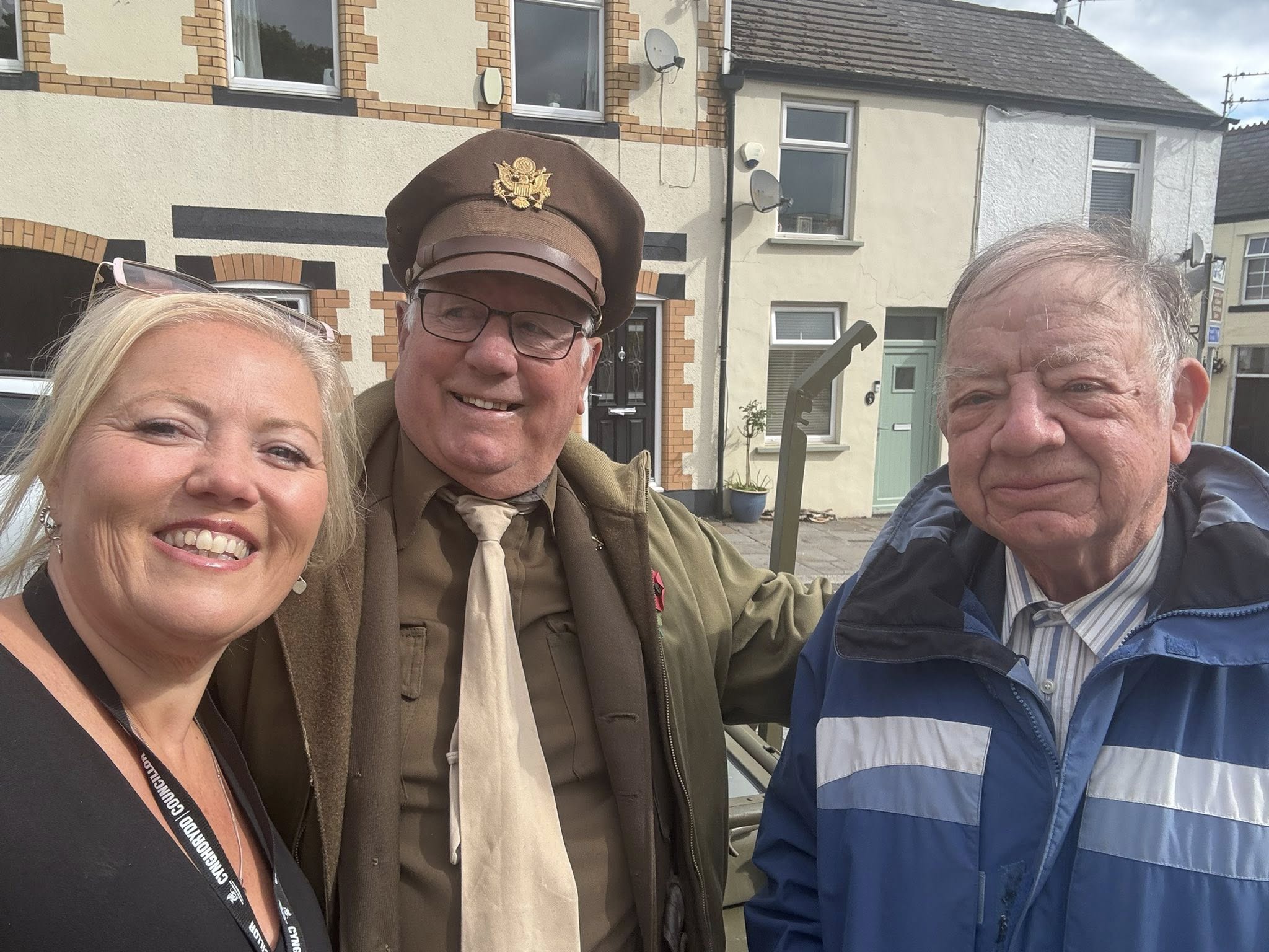 Trivallis Housing Landlord Wales Three people pose for a photo outdoors in front of houses. One man wears a vintage military uniform, the other man wears a blue jacket, and the woman in the foreground is smiling at the camera.