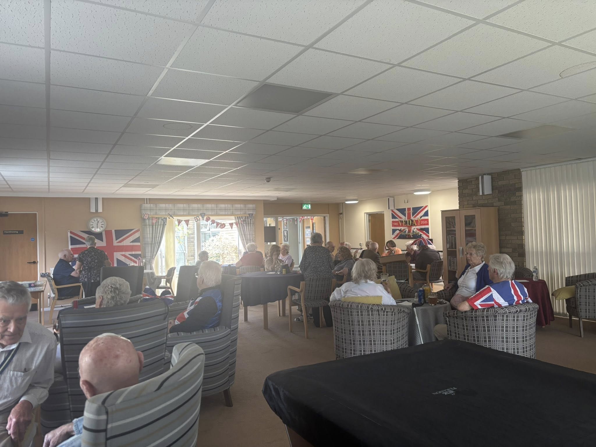 Trivallis Housing Landlord Wales A group of elderly people sit in armchairs in a communal room decorated with Union Jack flags. Some are chatting, while others face a window with a view outside. The room is well-lit with natural light.