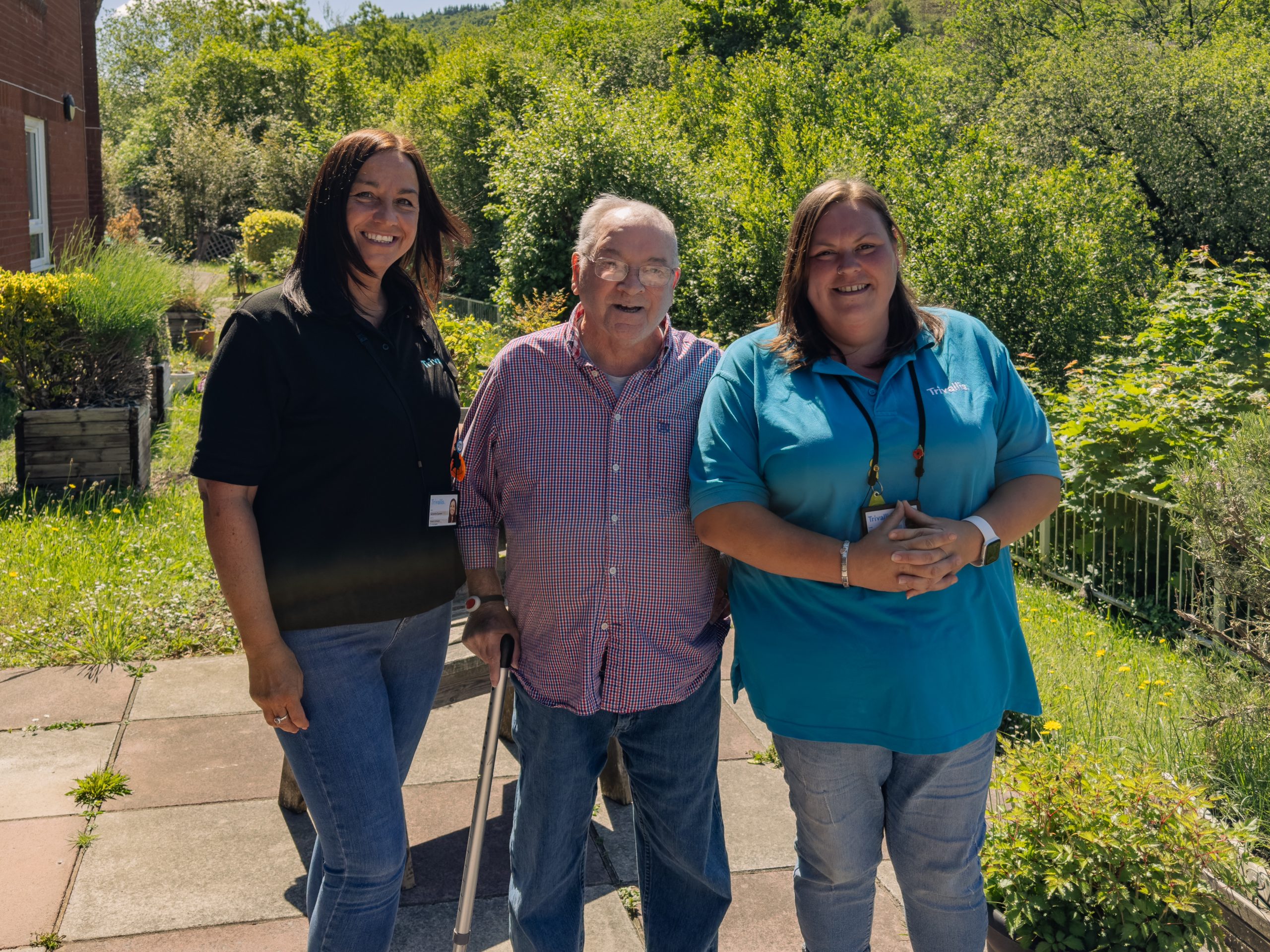 Trivallis Housing Landlord Wales Three adults stand outside on a sunny day. An older man with a walking stick stands between two women, all smiling at the camera. Trees and greenery fill the background.