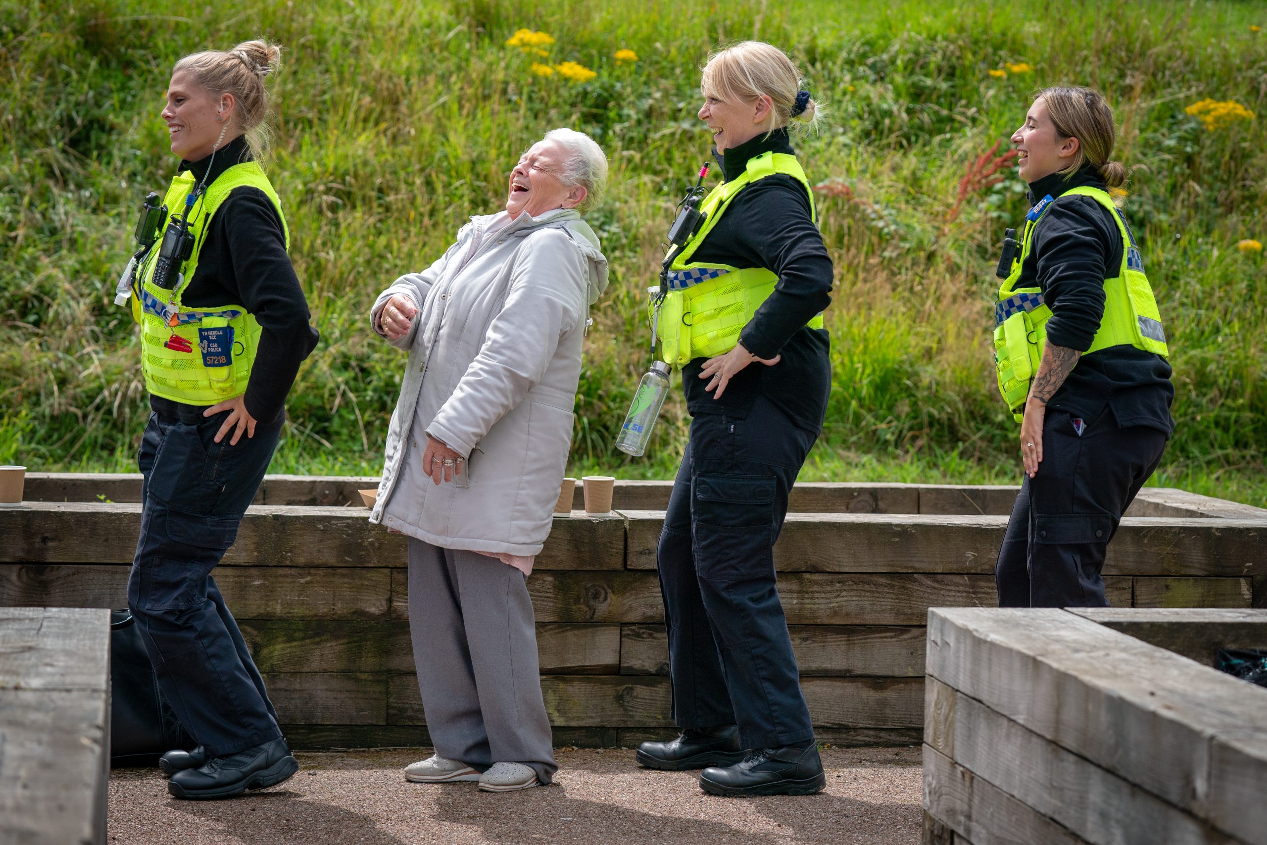 Trivallis Housing Landlord Wales An elderly woman and three police officers in yellow vests stand in a line outdoors, smiling and laughing, with hands on their hips in a lighthearted moment. Green grass and wooden barriers are in the background.