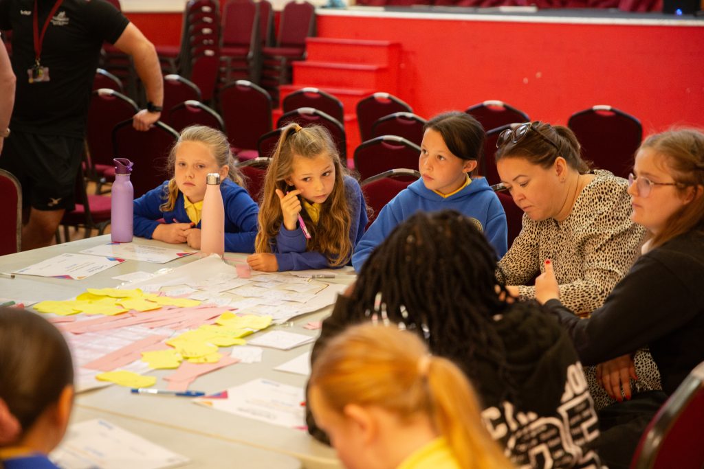 Trivallis Housing Landlord Wales A group of children and two adults sit around a table covered with papers and sticky notes, engaged in discussion. Rows of empty red chairs are visible in the background.