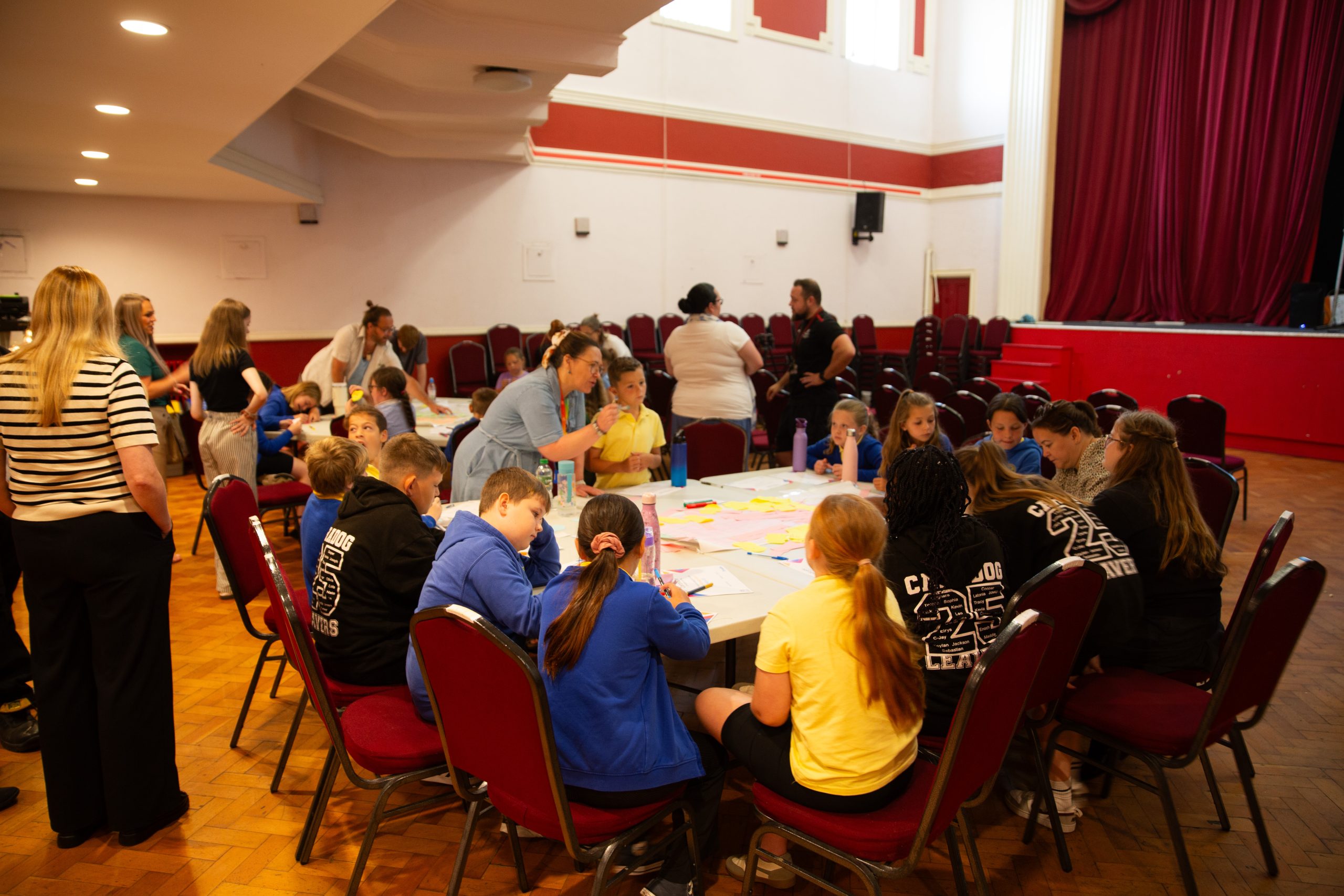 Trivallis Housing Landlord Wales A group of children and adults are gathered around a table in a large room, engaged in arts and crafts activities. Some people stand and observe, while others sit and work on colorful papers. Red chairs and stage are visible.