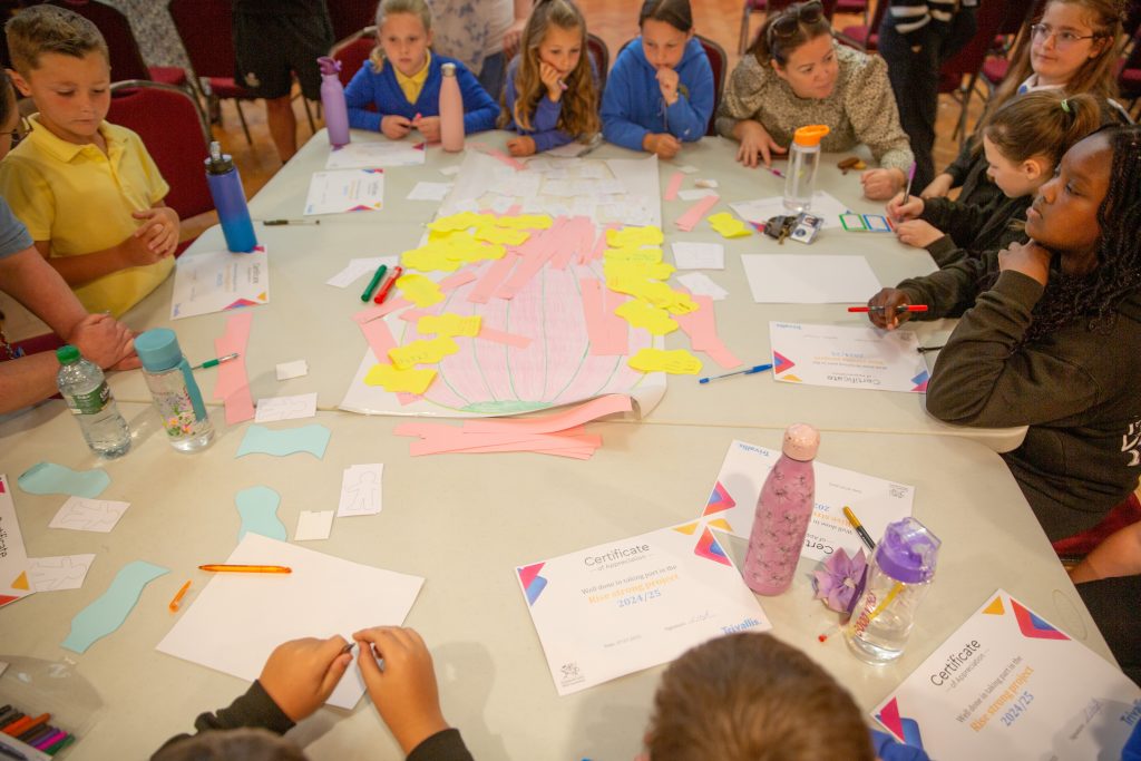 Trivallis Housing Landlord Wales A group of children and adults sit around a table covered with paper, pens, certificates, water bottles, and cut-out shapes, appearing to participate in an arts and crafts or group activity.