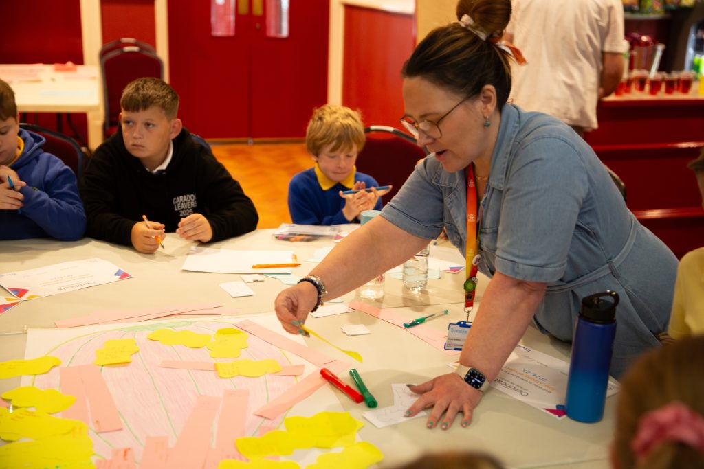 Trivallis Housing Landlord Wales A woman stands at a table covered with paper and yellow sticky notes, engaging a group of children seated around her. The children appear focused, writing with pens and markers. The background shows red chairs and doors.