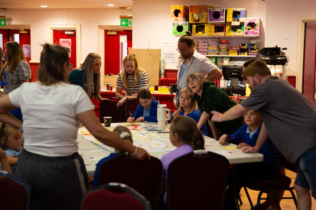 Trivallis Housing Landlord Wales A group of adults and children are gathered around a table covered with papers and sticky notes in a brightly lit room. The adults appear to be guiding or assisting the children with an activity.