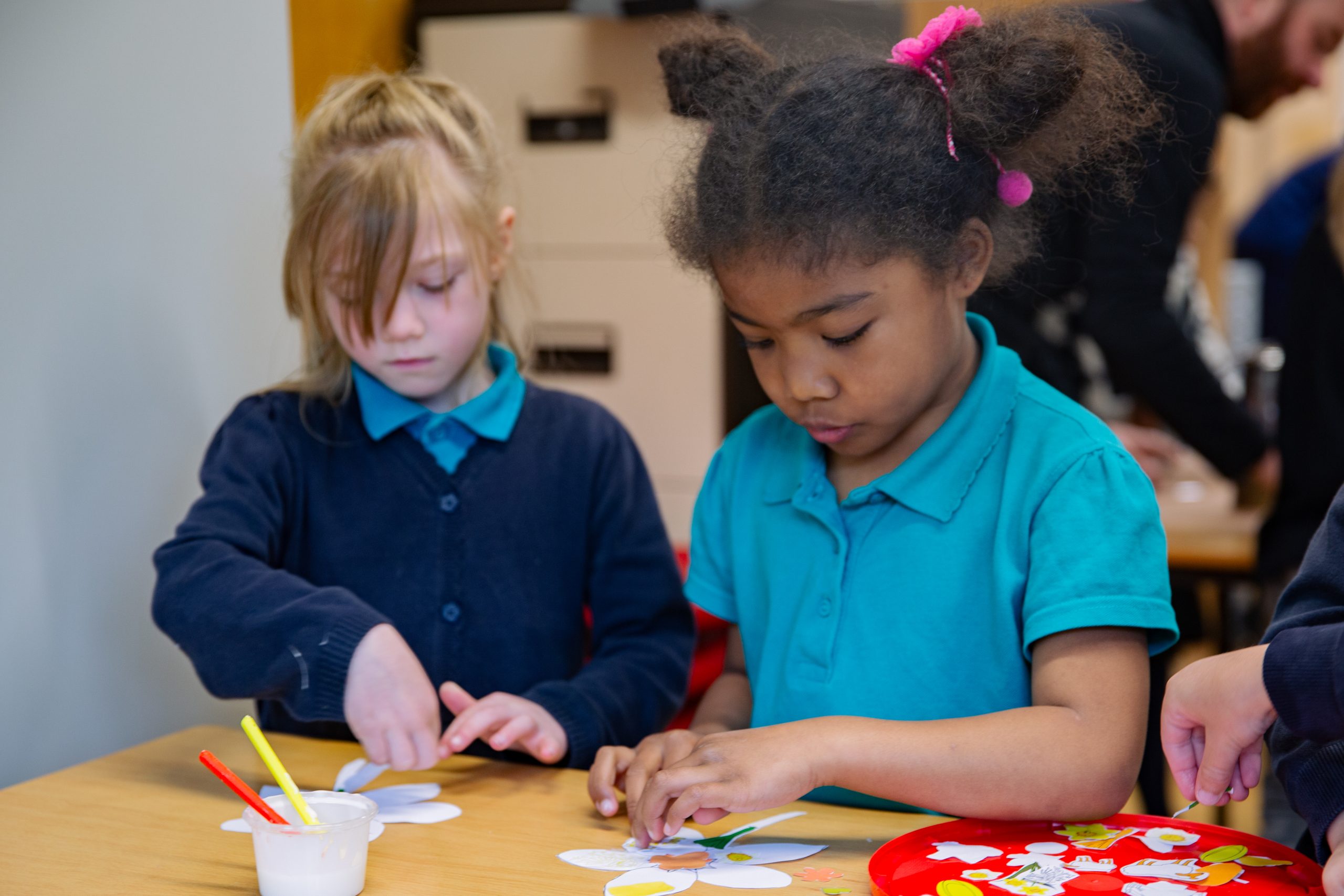 Trivallis Housing Landlord Wales Two young girls sitting at a table focus on a craft activity, gluing white paper flower shapes. Art supplies, including paintbrushes and glue, are on the table. A person is visible in the background.