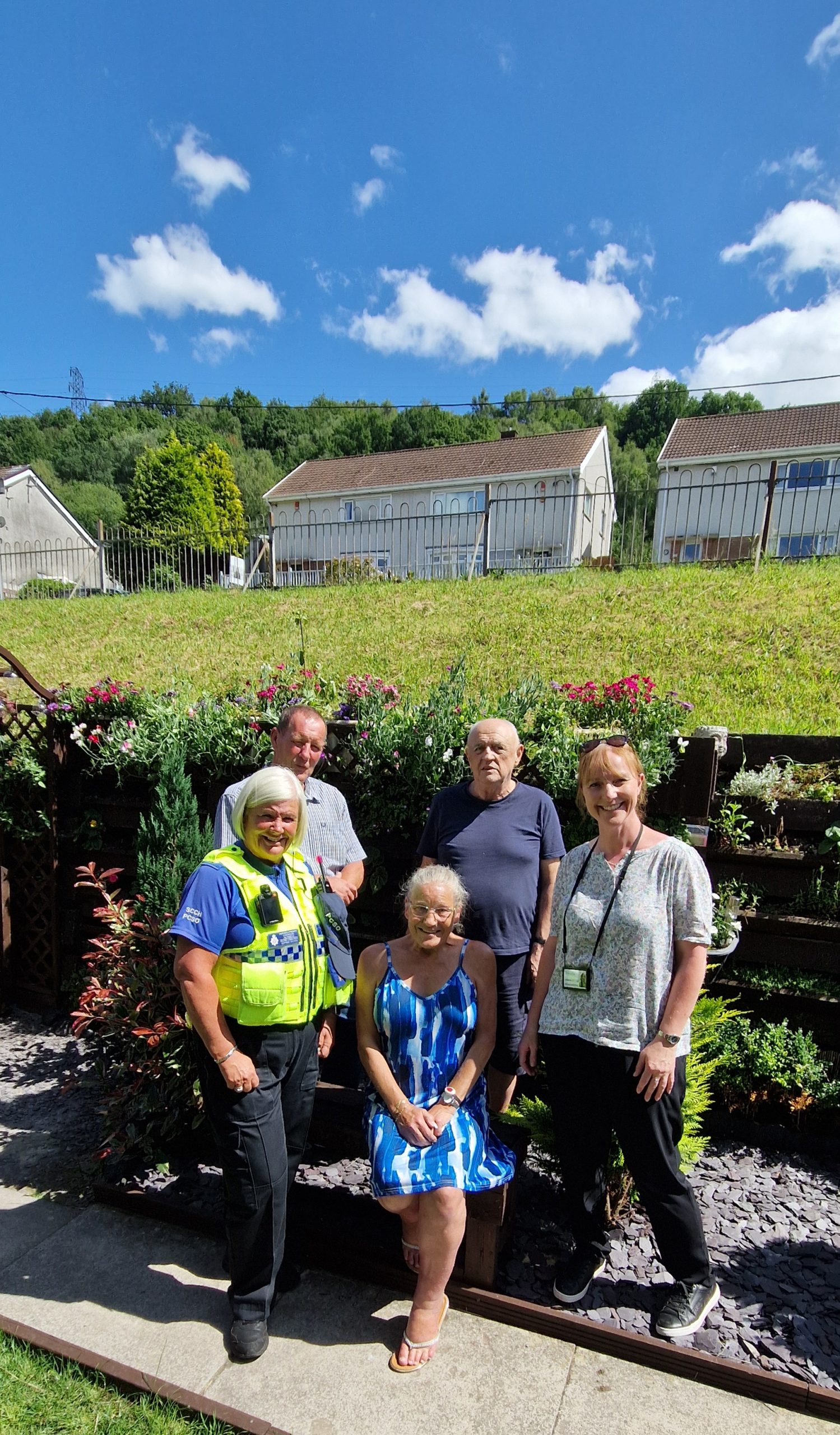 Trivallis Housing Landlord Wales Five adults, including a uniformed police officer, stand and kneel on a garden path in front of a raised flower bed, with green grass, houses, and a blue sky with scattered clouds in the background.