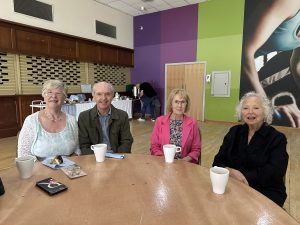 Trivallis Housing Landlord Wales Four older adults sit at a round table with white mugs, facing the camera. A woman stands at a buffet table in the background. The room has colorful walls and wooden accents.