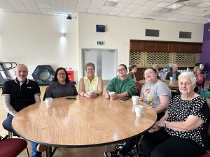 Trivallis Housing Landlord Wales Six people sit around a round wooden table with cups, smiling at the camera in a community hall setting. Other people are seated at tables in the background. The room has a high ceiling and bright lighting.
