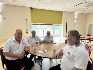 Trivallis Housing Landlord Wales Four men in white shirts sit around a round table with cups and a brochure in a spacious, bright room with wooden floors, large windows, and several empty red chairs.