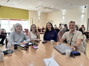 Trivallis Housing Landlord Wales Four adults sit at a wooden table in a brightly lit room with red chairs and other people in the background. Laptops, drinks, and notepads are on the table. All four look toward the camera and are smiling.