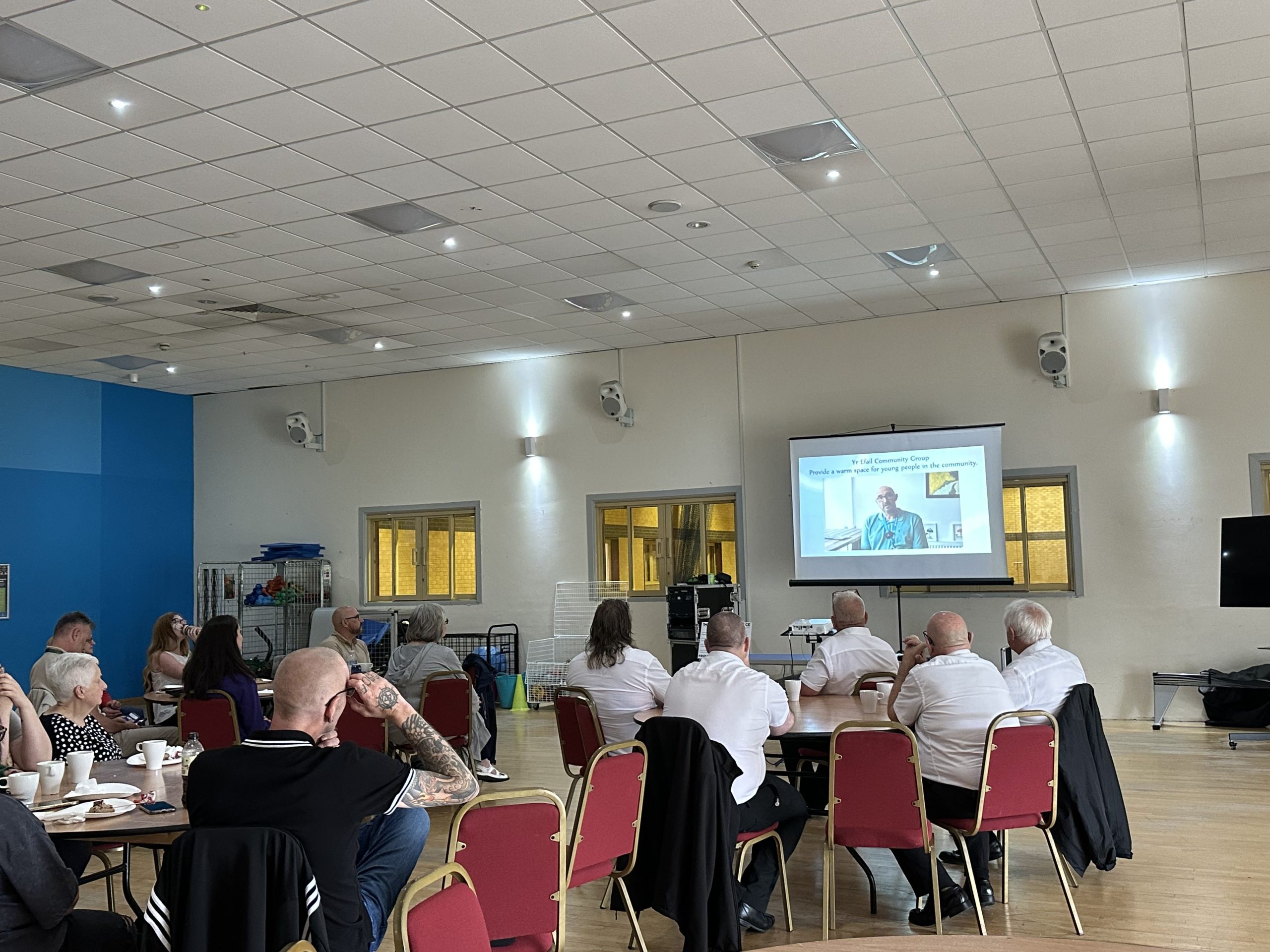 Trivallis Housing Landlord Wales People seated at round tables in a community hall watch a presentation projected on a screen at the front of the room. The audience faces forward, and the setting appears to be casual and professional.