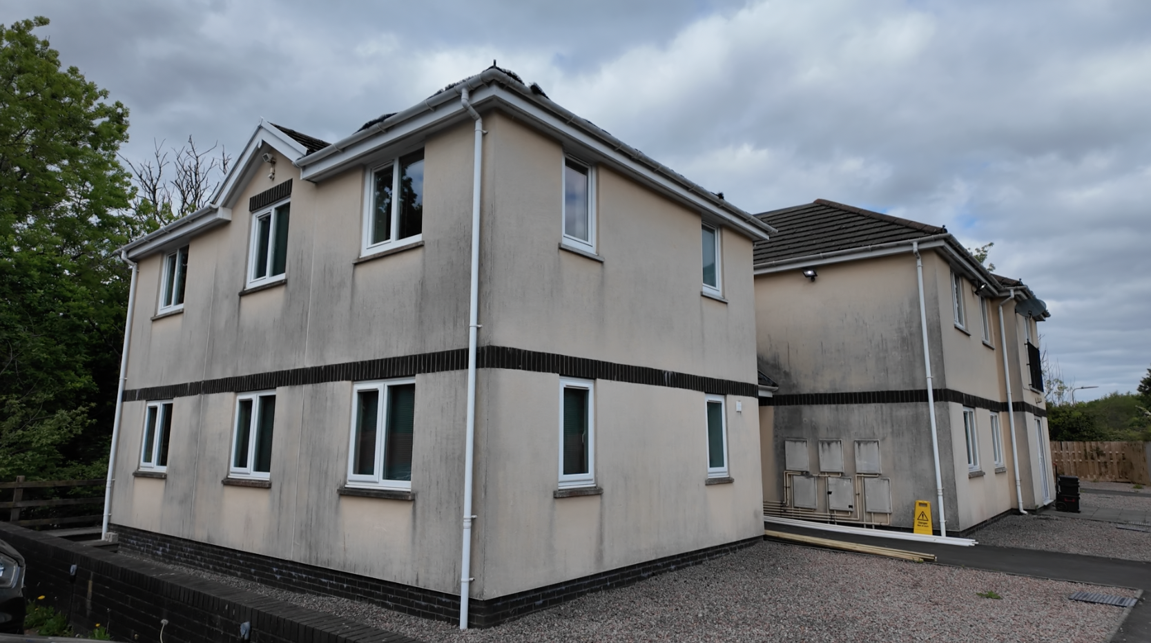 Trivallis Housing Landlord Wales Two cream-colored two-story buildings with black trim showing visible dirt and dark streaks on the walls, under a cloudy sky, with a gravel area and some wooden planks near the entrance.