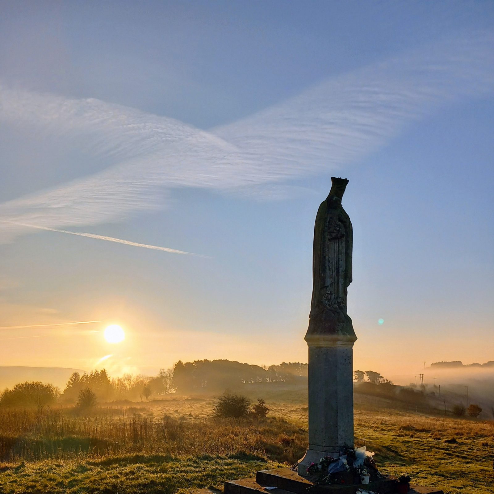 Trivallis Housing Landlord Wales Our Lady of Penrhys, a statue on a pedestal stands in a grassy field at sunrise, with the sun low on the horizon and mist in the background. Some flowers and items are placed at the base of the statue. Trees are visible in the distance.