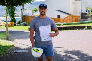 Trivallis Housing Landlord Wales A man in a gray shirt and shorts stands outdoors holding a white hard hat and a certificate. There are houses, trees, and a wooden fence in the background.