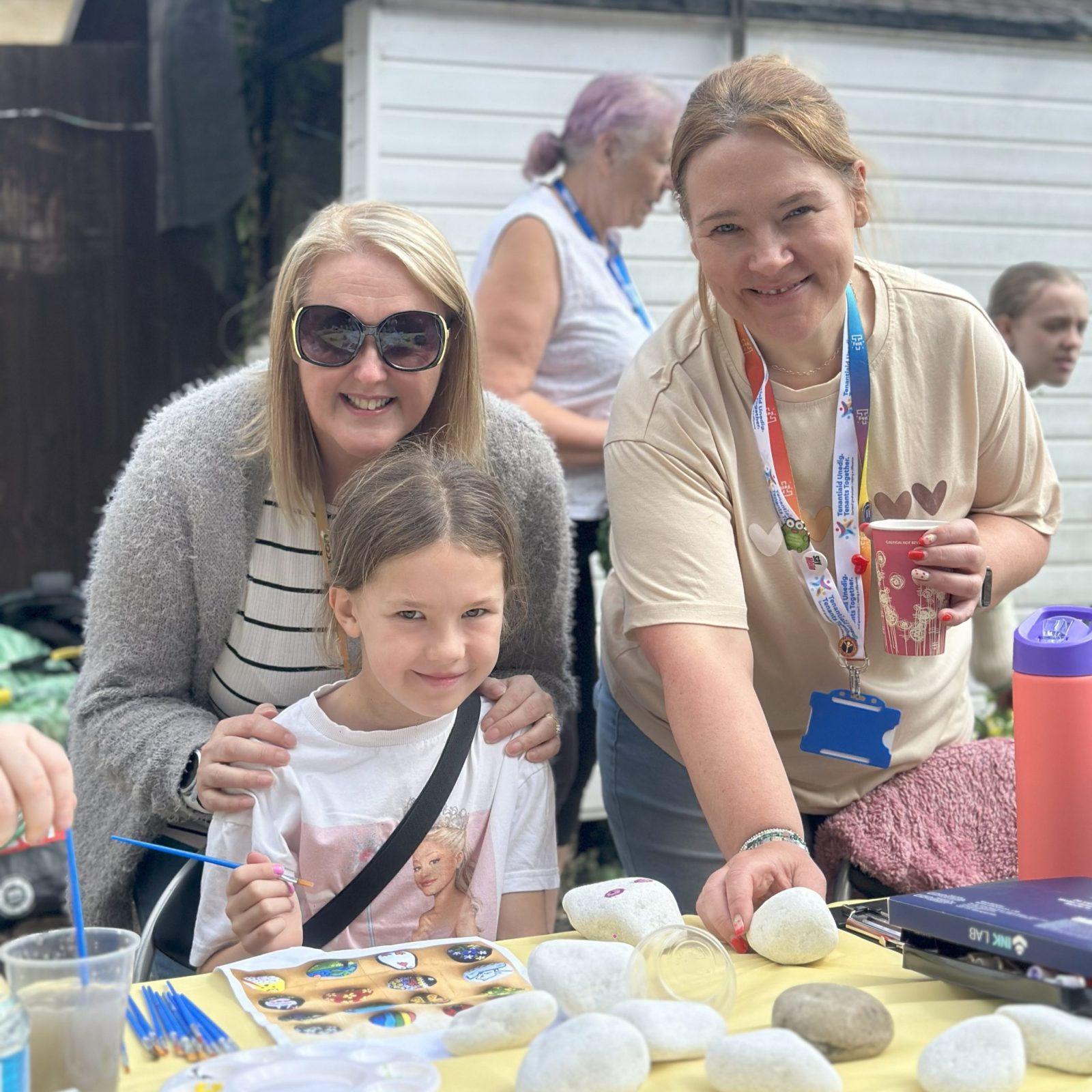 Trivallis Housing Landlord Wales A woman and a girl pose and smile at a table covered with white stones, paints, and brushes. Another woman with a lanyard stands nearby holding a cup. Other people are visible in the outdoor background.