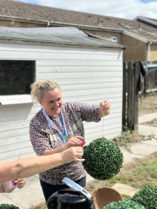 Trivallis Housing Landlord Wales A woman smiling and holding a string attached to a round artificial topiary ball outdoors, with another hand assisting; a white shed and houses are visible in the background.