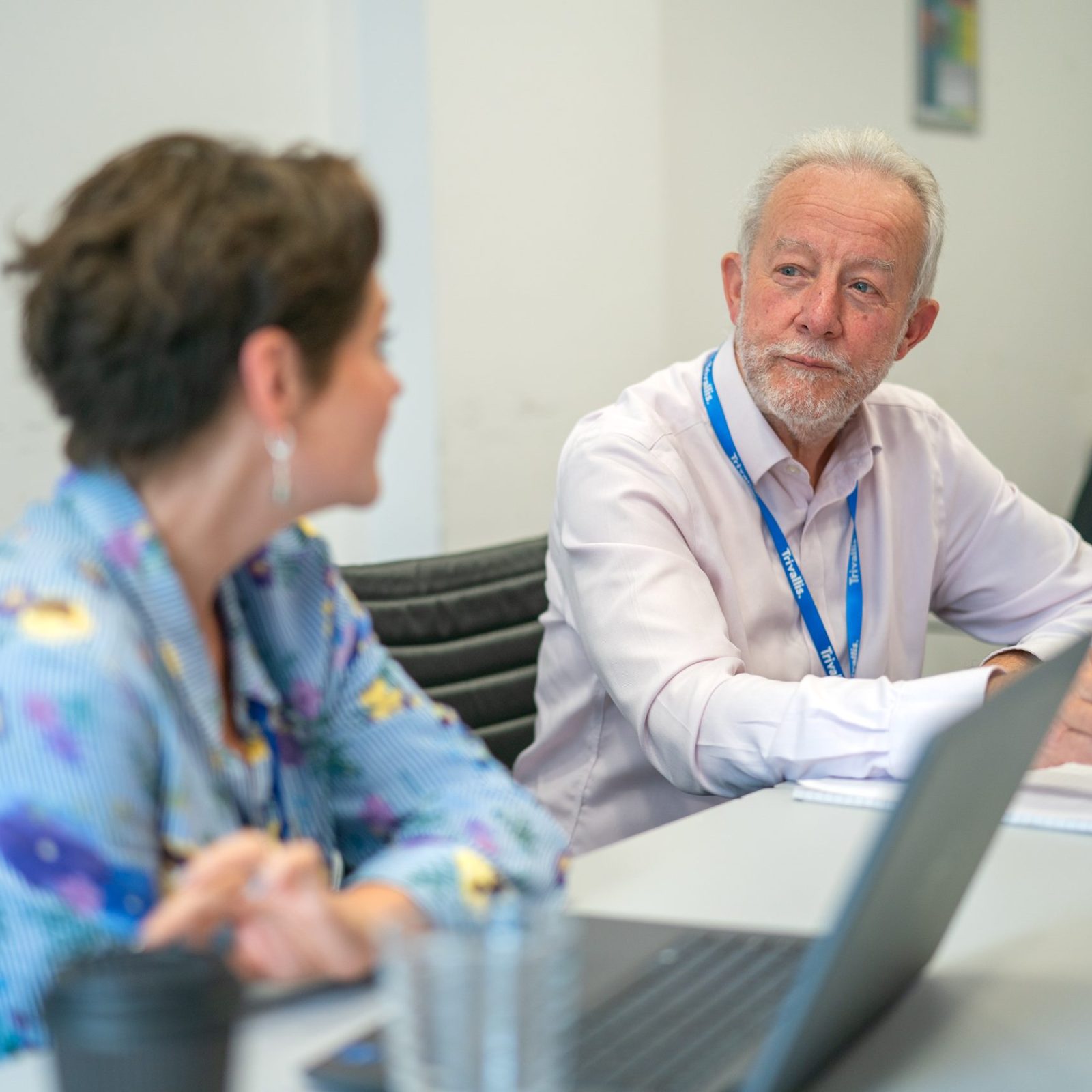 Trivallis Housing Landlord Wales Two people sit at a table with laptops and notepads, having a conversation in a meeting room. The man is looking attentively at the woman, who is slightly out of focus in the foreground.