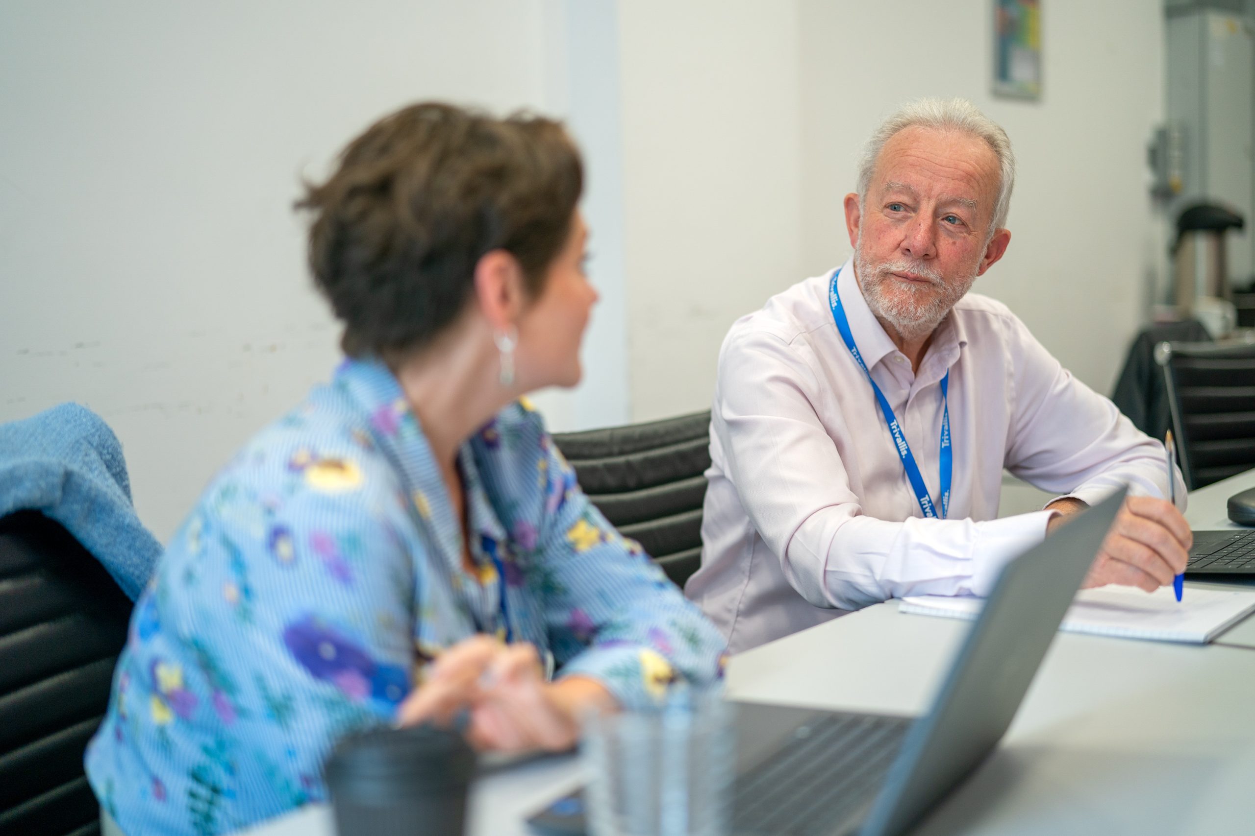 Trivallis Housing Landlord Wales Two people sit at a table with laptops and notepads, having a conversation in a meeting room. The man is looking attentively at the woman, who is slightly out of focus in the foreground.