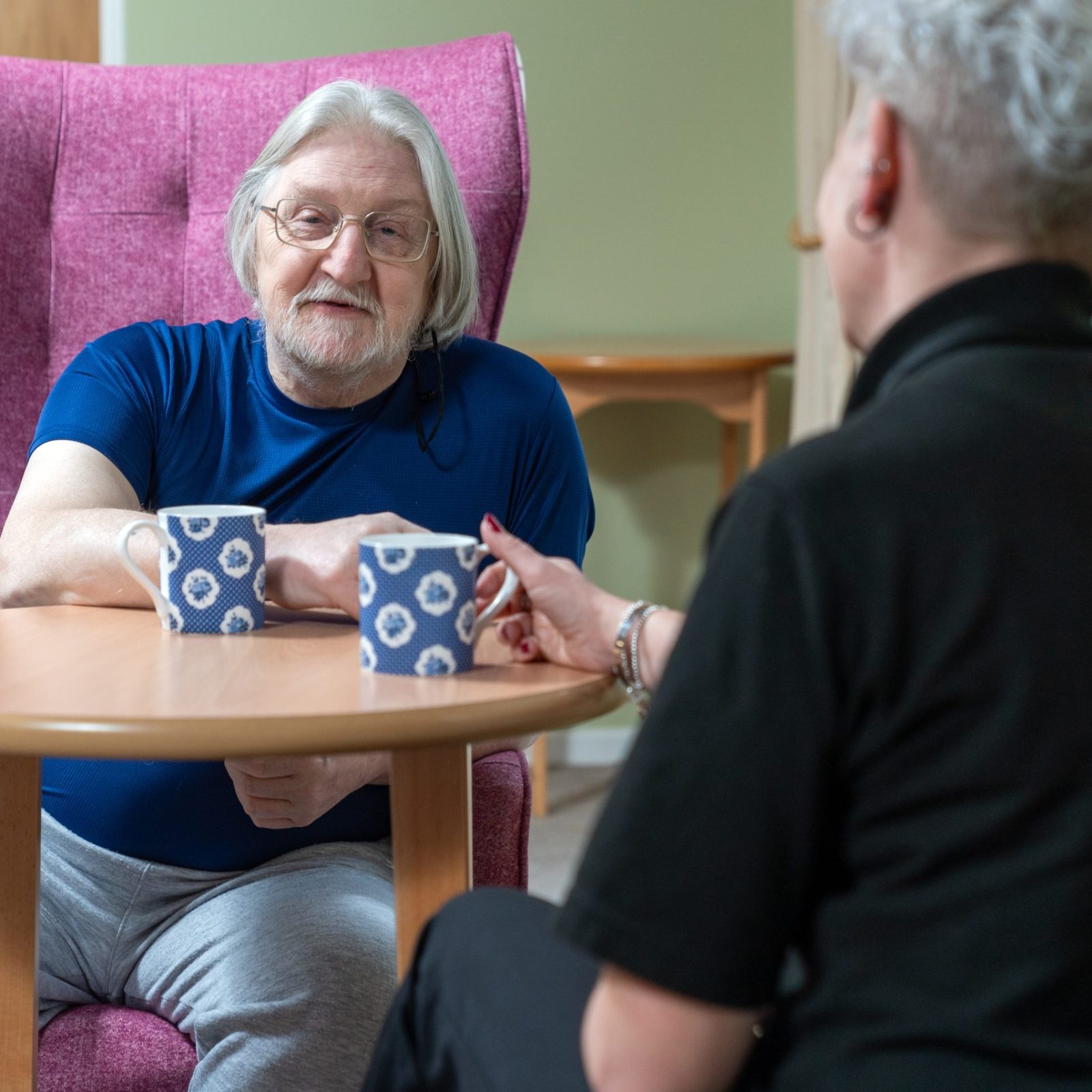 Trivallis Housing Landlord Wales An older man with gray hair and glasses sits in a pink chair across from another person at a small round table. Both have mugs with a blue and white pattern. The setting appears to be indoors, possibly in a care facility.