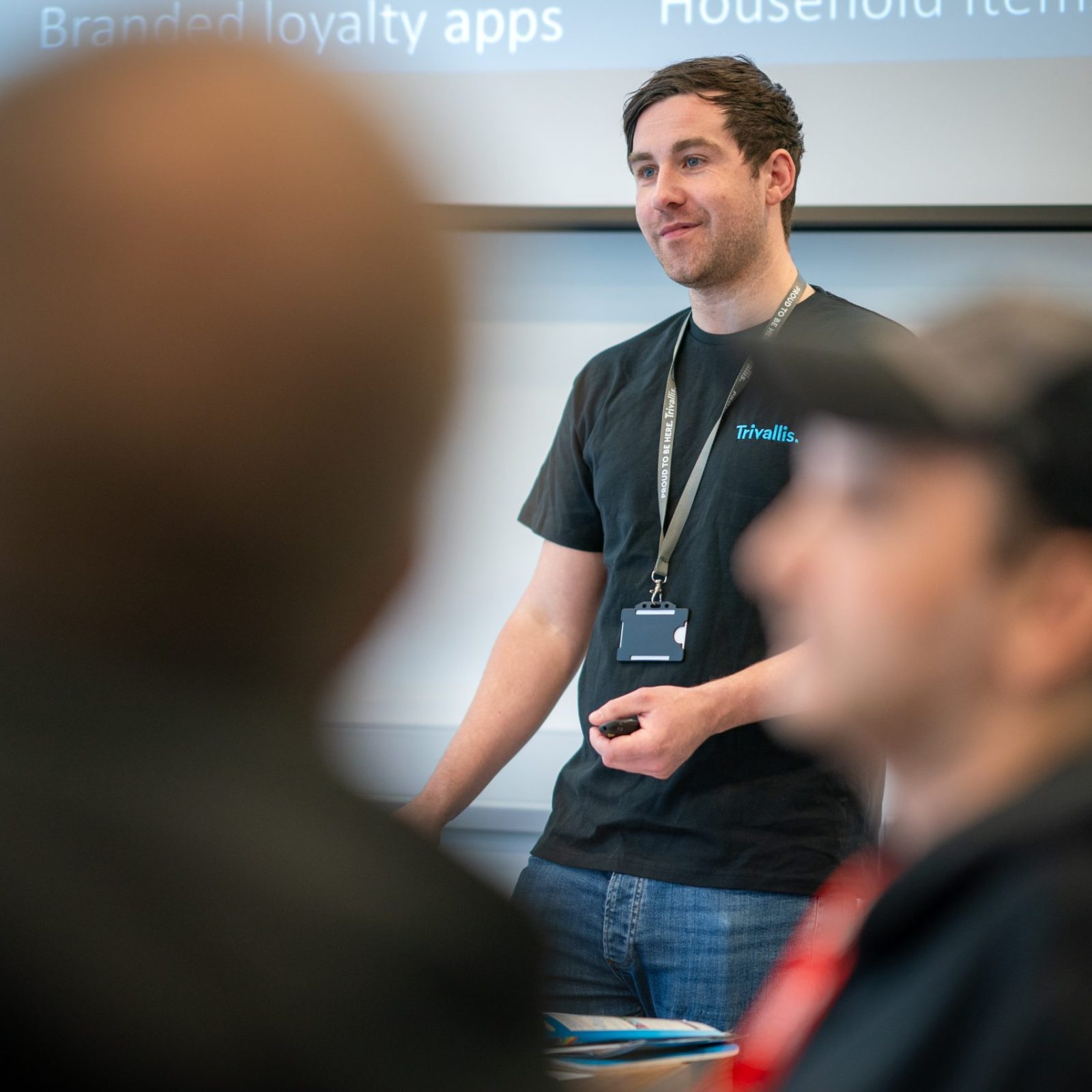 Trivallis Housing Landlord Wales A man stands at the front of a room giving a presentation, with a screen behind him displaying partial text. Two people in the foreground are out of focus, listening to the presenter.