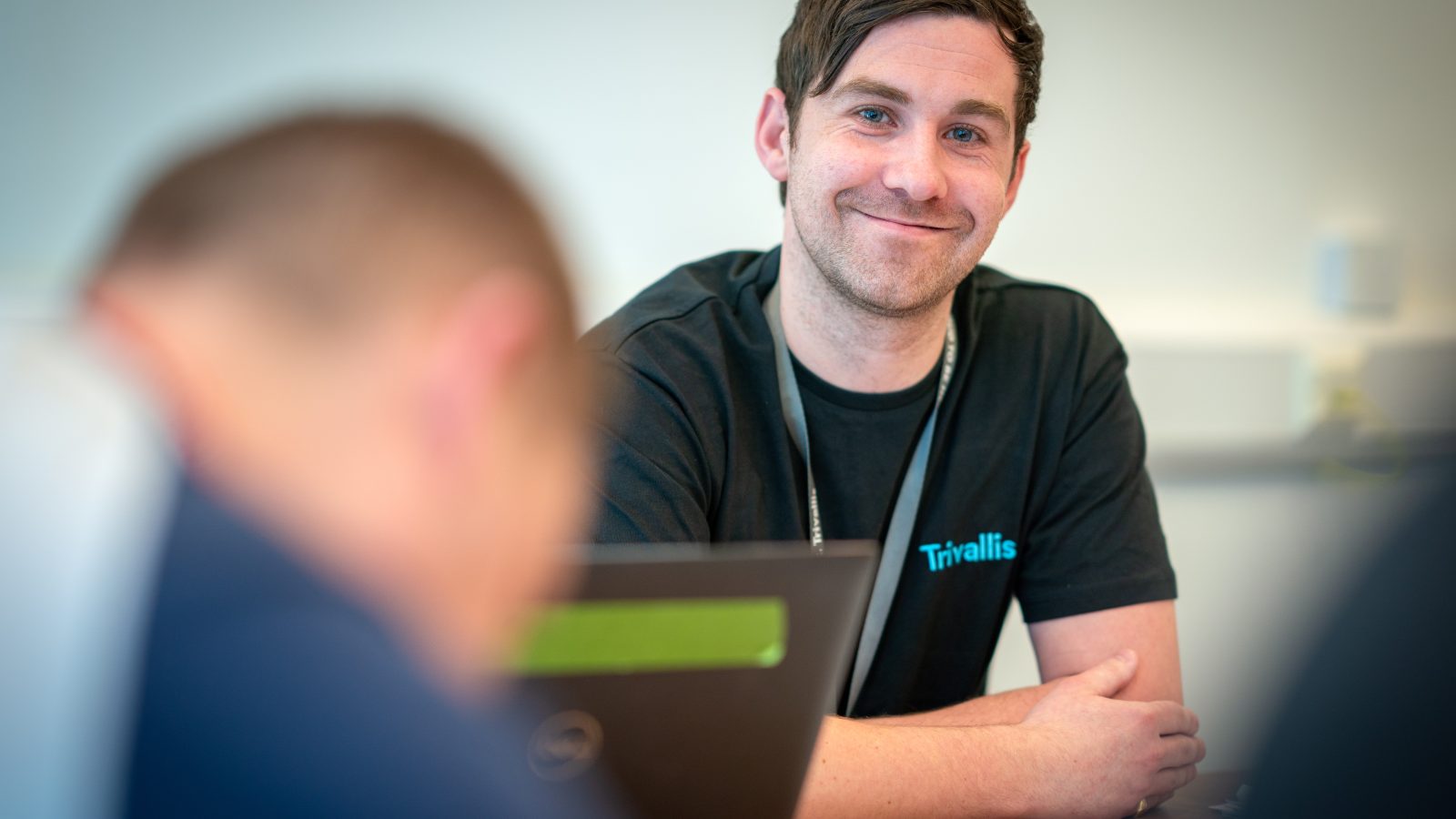 Trivallis Housing Landlord Wales A man wearing a black Trivallis shirt sits at a table, smiling at the camera. A laptop and papers are in front of him. Another person is blurred in the foreground. The setting appears to be an office or meeting room.