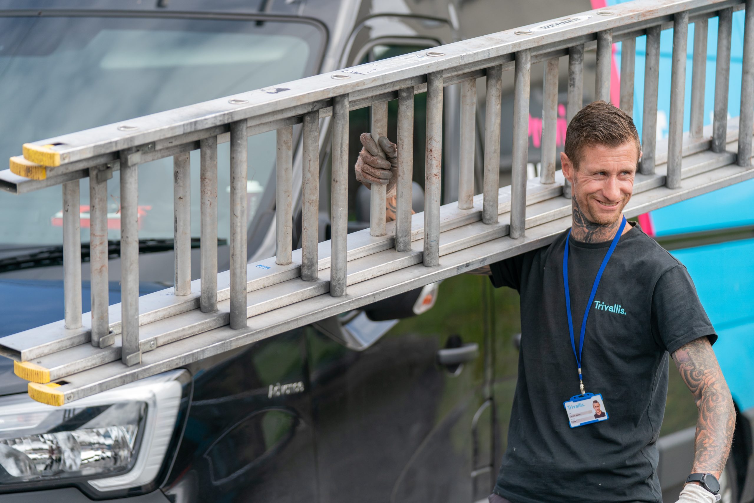 Trivallis Housing Landlord Wales A man with tattoos wearing a black t-shirt and a lanyard carries a long metal ladder on his shoulder, standing in front of a parked van.
