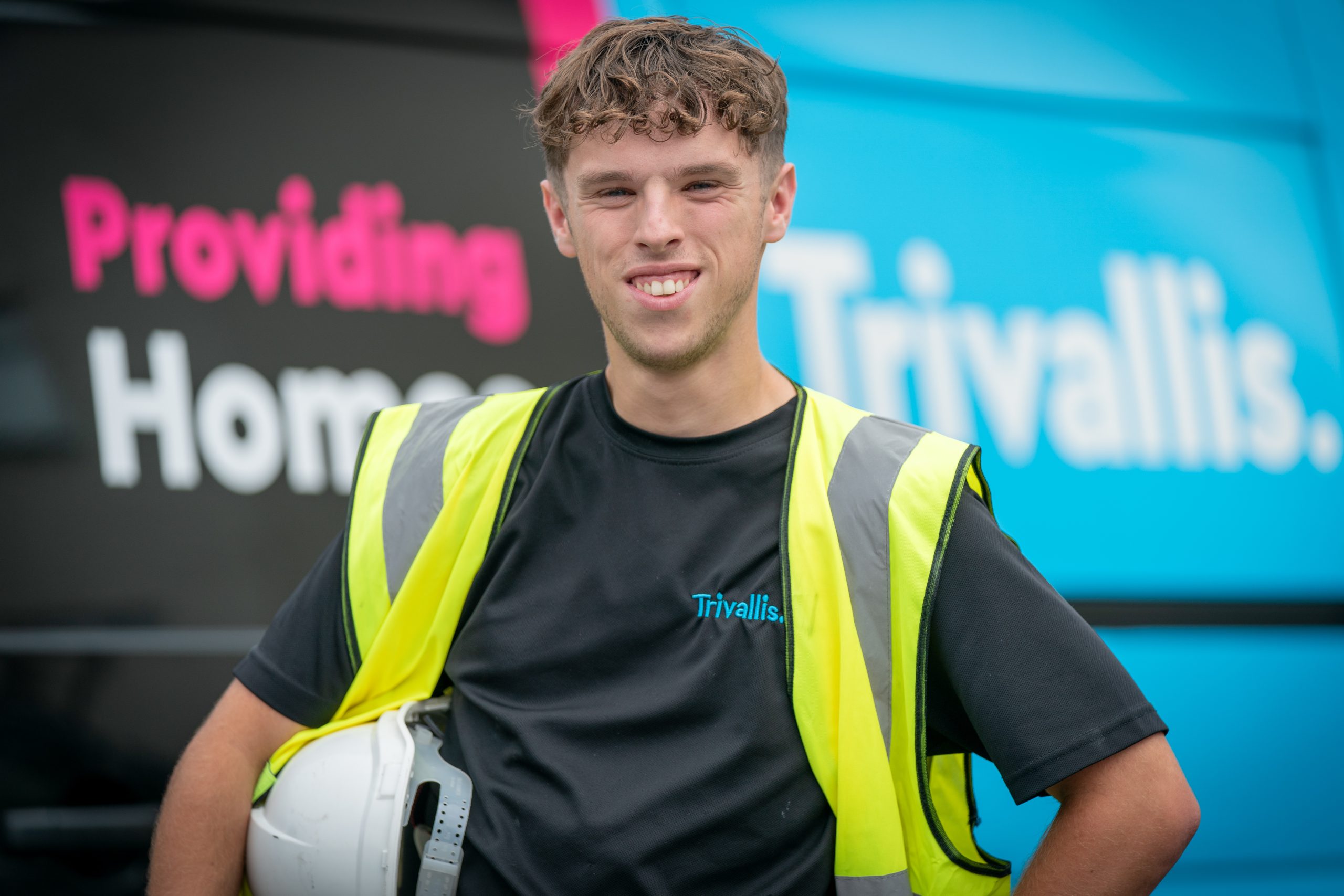 Trivallis Housing Landlord Wales A young man wearing a yellow safety vest and holding a white hard hat stands in front of a van with the "Trivallis" logo and the words "Providing Homes" partially visible in the background.