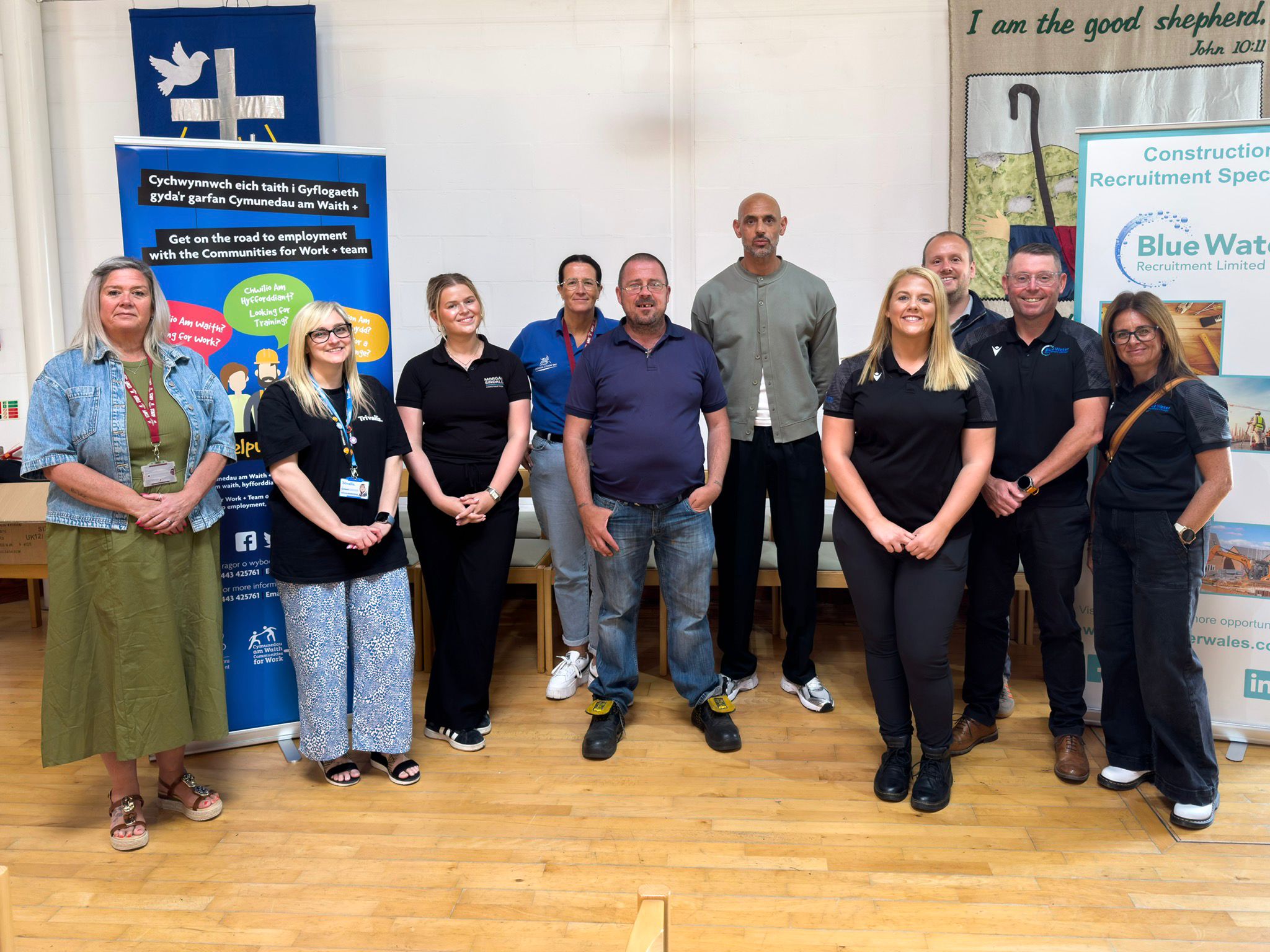 Trivallis Housing Landlord Wales Ten adults stand indoors on a wooden floor, posing for a group photo. Two roll-up banners about employment opportunities are visible on either side. The wall behind them features a mural and a sign that reads “I am the good shepherd.”.