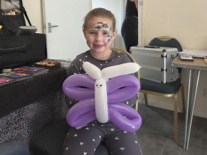 Trivallis Housing Landlord Wales A young girl with face paint sits indoors, smiling and holding a balloon animal shaped like a butterfly with white and purple sections. She is wearing a grey outfit with white patterns.